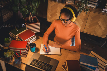 Young woman studying indoors with headphones, surrounded by books and laptop, creating a cozy and...