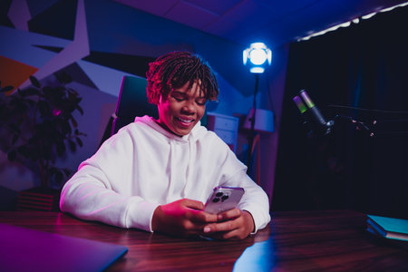 Positive man in a neon-lit room using smartphone while podcasting with microphone showcasing...