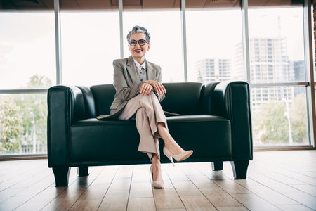 Confident mature businesswoman in elegant suit seated on a stylish sofa in modern office workspace with city viewの写真素材
