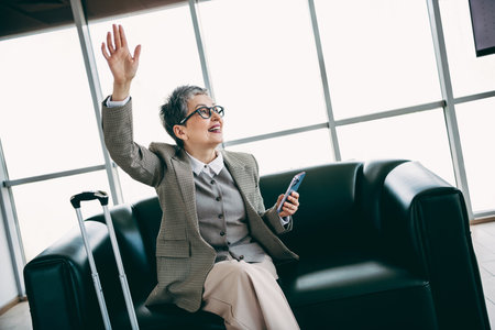 Elegant senior businesswoman waving in office lounge using smartphone emphasizing confidence and professional attireの写真素材