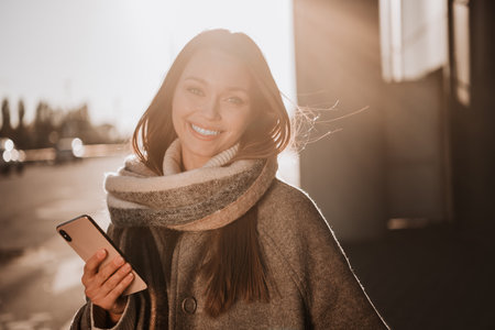Outdoor portrait of a young woman in autumn setting holding phone and smiling in sunlightの写真素材