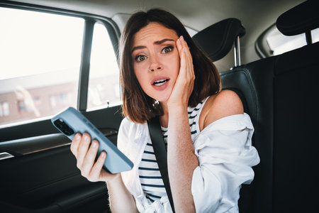 Young woman surprised in car, holding smartphone, wearing casual clothes, experiencing city sights on a sunny dayの写真素材