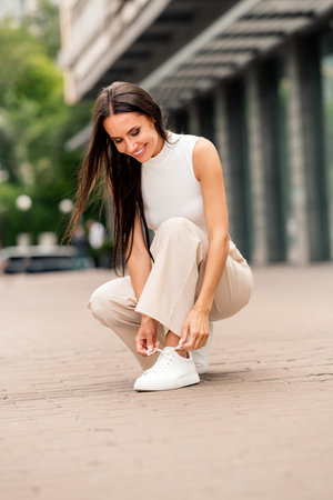 Young stylish woman outdoors tying shoes, showcasing fashionable attire in a sunny urban setting with confidence and eleganceの写真素材