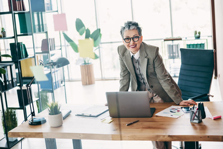 Confident businesswoman standing by office desk with professional attire surrounded by modern workspace elements in a corporate environmentの写真素材