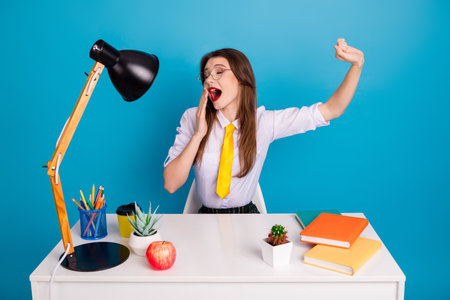 Young woman yawning at her desk surrounded by book, stationery, and plants against a vibrant blue backgroundの写真素材