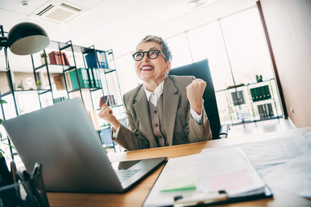 Confident businesswoman celebrating success at her office desk in bright corporate surroundingsの写真素材