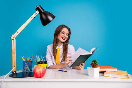 Smart woman in retro style sitting with study materials and lamp, cheerful and engaged in education, blue backdropの写真素材