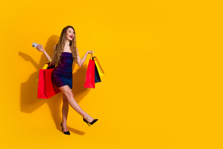 Fashionable young woman carrying colorful shopping bags while holding a phone and smiling against a yellow backgroundの写真素材