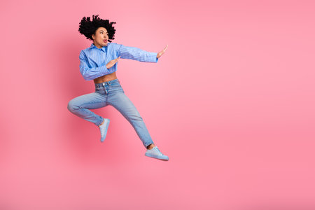 Excited young woman jumping mid-air in casual outfit with pink background showing energetic gesture and movementの写真素材