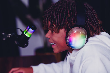 Young gamer streaming with neon-lit headphones using a microphone in a casual indoor setup during the eveningの写真素材