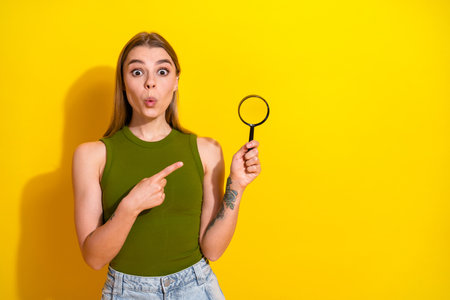 Adorable young woman poses with a magnifying glass on a vibrant yellow background, expressing surprise and charm in casual fashionの写真素材