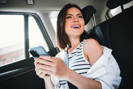 Young woman enjoying a sunny day, smiling inside a car, wearing a stylish striped shirt and holding a smartphone while traveling.の写真素材