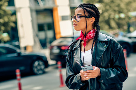 Young stylish woman in a leather jacket holding a smartphone while walking outdoors on a sunny urban...