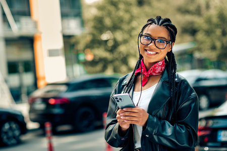 Lovely woman enjoying a day out in the city, wearing a stylish leather jacket and scarf, holding her smartphone outdoorsの写真素材
