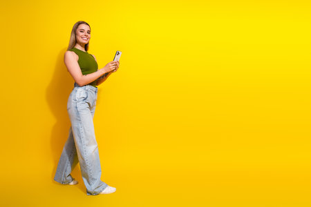 Young woman in green top and jeans holding a smartphone, smiling cheerfully, standing against a bright yellow backgroundの写真素材