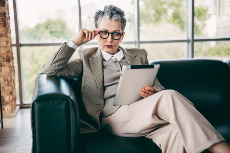Mature businesswoman in formal attire working on a tablet device while seated in a professional office environmentの写真素材