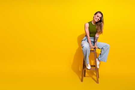 Young woman smiling while sitting on a chair against a vibrant yellow background wearing trendy casual attireの写真素材