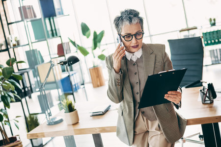 Mature businesswoman in office using phone and reviewing documents, showcasing confidence, professional style, and an elegant workspaceの写真素材
