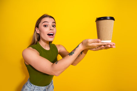 Excited young woman holding a cup and smiling against a vibrant yellow background while wearing a green casual topの写真素材