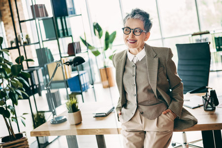 Experienced businesswoman in stylish attire smiling confidently while leaning on a desk in a modern office setting with bright decorの写真素材