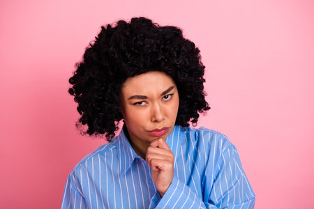 Minded woman with curly hair posing against a pink background in a thoughtful expression while...