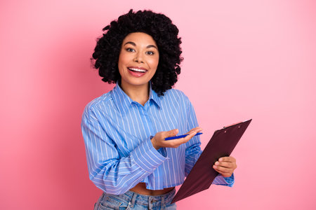Confident young woman holding a clipboard while smiling brightly and gesturing against a pink background showcasing positivityの写真素材