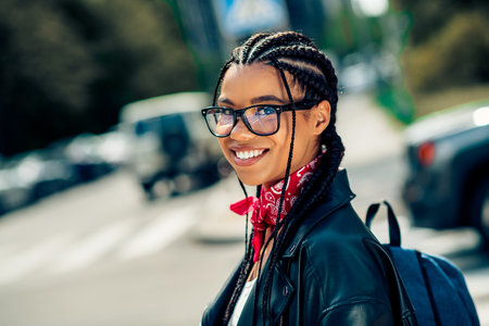 Young woman with braided hair wearing a leather jacket and glasses, confidently smiling on a sunny day in the cityの写真素材