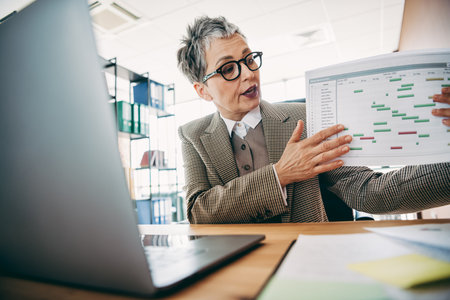 Confident professional pointing to chart while explaining business strategy during office meeting with modern workspace settingの写真素材