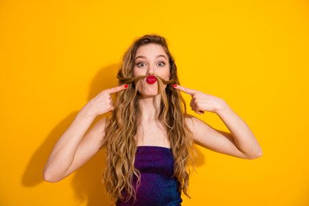 Playful blonde woman making quirky gesture with hair and red lips on yellow background, showcasing fun fashion styleの写真素材