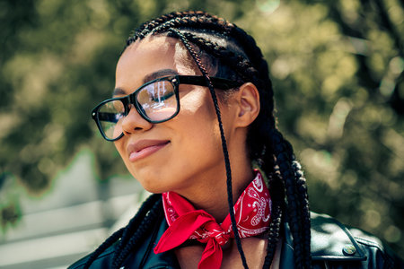 Young woman with braids and glasses outdoors, wearing a leather jacket and scarf, radiating charm and casual eleganceの写真素材