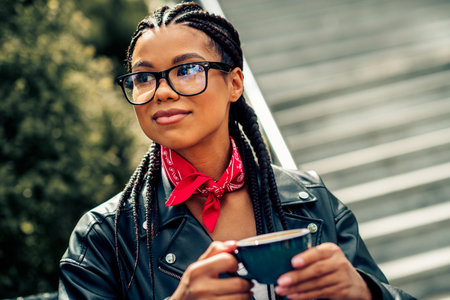 Lovely woman with braided hair and glasses enjoying a hot drink outdoors on a sunny day in casual leather jacketの写真素材