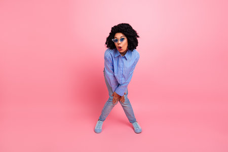 Young stylish woman with curly hair posing playfully against pink background wearing blue striped shirt and jeansの写真素材
