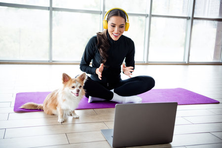 Charming woman practicing indoor workout with laptop and her dog in stylish activewear, enjoying a...