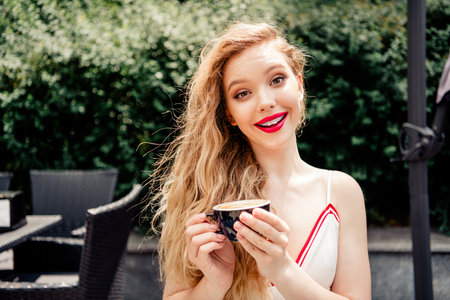Young woman enjoying coffee outdoors in a summer setting with a cheerful expression and stylish appearanceの写真素材