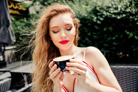 Young woman enjoying a cup of coffee outdoors in a park, showcasing fashion, elegance, and relaxation on a sunny day.の写真素材