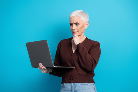 Mature woman in casual attire thinking while using a laptop isolated on a bright blue background for...