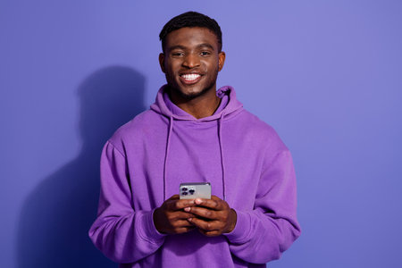 Young man in purple hoodie holding smartphone and smiling on violet background promoting casual lifestyleの写真素材