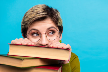 Young woman with short hair and glasses poses charmingly with book against a vibrant blue backgroundの写真素材