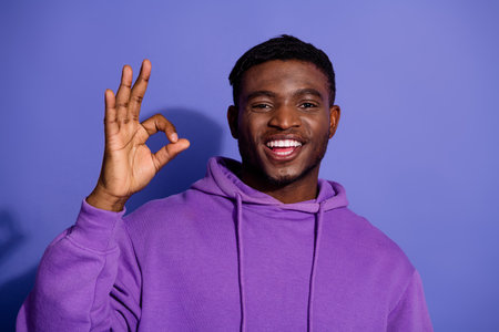 Smiling young man in casual purple hoodie showing OK gesture against vivid wall for modern lifestyle promotionsの写真素材