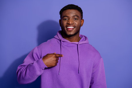 Young smiling man in a purple hoodie pointing confidently against a vibrant violet backgroundの写真素材