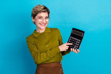 Smiling young woman in casual khaki shirt holding a calculator on blue background emphasizing professional and educational themesの写真素材
