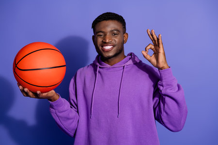Young man in purple hoodie holding a basketball and making an OK hand gesture against a violet backgroundの写真素材