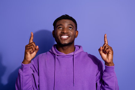 Portrait of a cheerful young man in a purple hoodie pointing upwards against a vibrant violet...