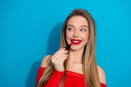 Blonde woman in red top holding lipstick against blue background and smiling with a vibrant and happy expressionの写真素材