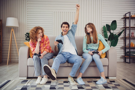 Funny young people enjoying video gaming on the couch, expressing joy and concentration in a cozy living room setting with a relaxed vibeの写真素材