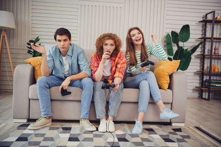 Three young friends enjoying video games together on a cozy living room sofa during a relaxing...