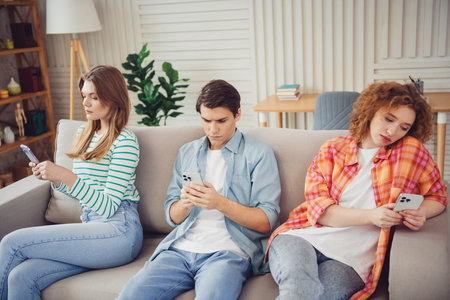 Funny young friends sitting on a sofa using smartphones in a cozy living room, depicting a casual...