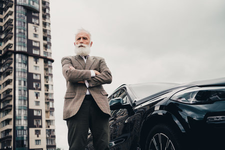 Handsome mature businessman standing beside a car near a modern building in an urban setting on a cloudy dayの写真素材