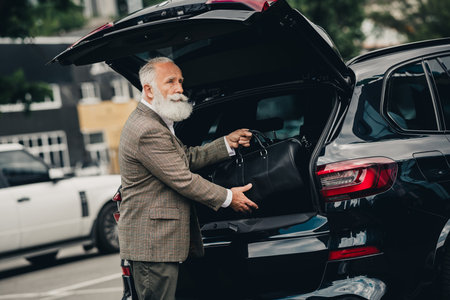 Senior businessman with stylish jacket loading suitcase into luxury car in an urban environment on a...