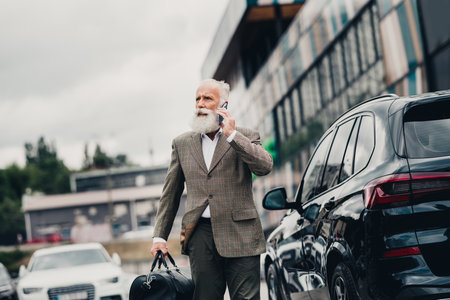 Handsome businessman confidently walking outdoors with stylish formal attire and a travel bag while talking on phoneの写真素材